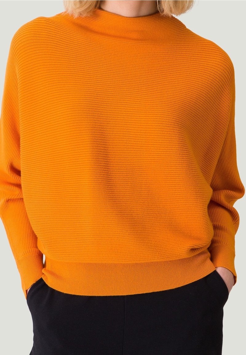 Personne portant un pull en maille côtelée orange vif à manches longues et un pantalon noir, les mains dans les poches, sur un fond clair.
