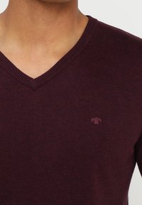 V-Ausschnitt-Pullover aus dunkelburgunderfarbenem Strickstoff. Mit einem dezenten Logo-Detail auf der Brust. Weiche Textur mit einer figurbetonten Silhouette.