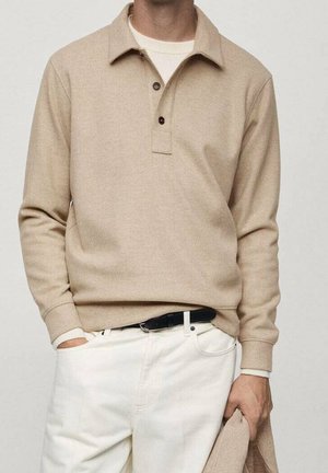 Homme portant un polo beige à manches longues, un pantalon blanc avec une ceinture noire, tenant un tissu beige assorti dans la main droite, debout devant un fond uni.