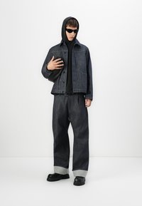 Mand iført mørk denimjakke og bukser med opsmøg, sort hoodie, solbriller, sorte sko og holder en sort taske mod en hvid baggrund.