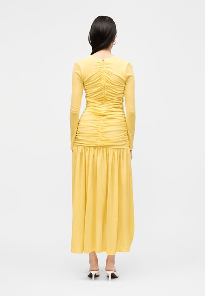 Femme aux cheveux noirs raides, portant une robe jaune froncée à manches longues et des mules blanches à talons, debout de dos sur un fond uni.