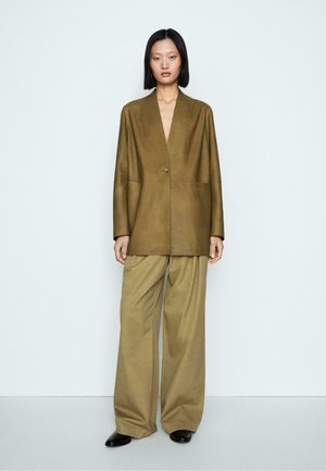 Femme aux cheveux noirs lisses porte un blazer oversize olive et un pantalon ample, debout devant un fond uni gris clair.