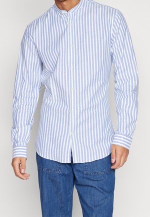 Man wearing a light blue and white vertical striped button-up shirt with long sleeves and blue denim pants.