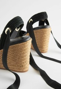 Sandali con zeppa intrecciata nera con base in corda di juta, dotati di cinturini in tessuto nero e dettagli in metallo dorato. Suola nera liscia.