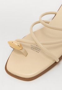 Beige flade sandal med tynde, krydsende remme og et guldblad-detalje på tæerne. Firkantet tåform; glat, matte tekstur.