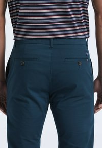 Blauwe katoenen broek met een soepele textuur, voorzien van twee achterzakken met knoopsluitingen en een klein merketiket dat aan de tailleband is vastgenaaid.