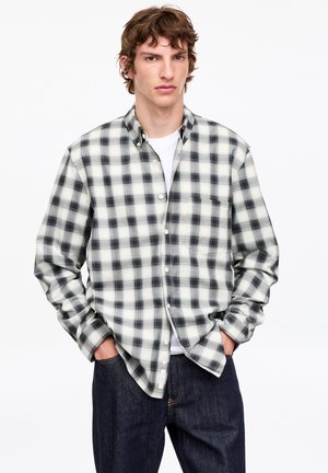 Jeune homme aux cheveux bruns ondulés portant une chemise à carreaux noire et blanche boutonnée et un jean bleu foncé, les mains dans les poches, faisant face.