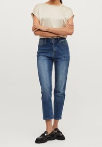 Haut beige en satin à manches courtes, jean en denim bleu taille haute, raccourci au niveau des chevilles, associé à des mocassins noirs avec des accents en métal doré.