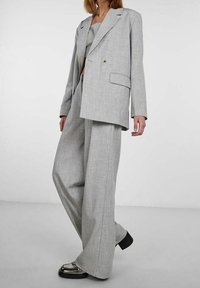 Jasnoszary garnitur na miarę z luźno dopasowaną marynarką i spodniami o szerokich nogawkach, wykonany z teksturowanej tkaniny z subtelnym wzorem i kontrastującymi przeszyciami.