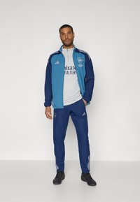 Herre sportstøj med en blå lynlåstrøje, der har hvide striber og logo, parret med matchende blå bukser og sorte sko.