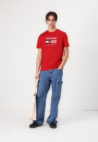 Červené bavlněné tričko s bílým logem 'HILFIGER' a designem vlajky. Spárované s modrými džínovými cargo kalhotami. Model drží béžovou tašku.