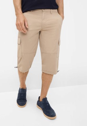 Mand iført beige cargoshorts med klaplommer og marineblå sneakers, der står mod en hvid baggrund.
