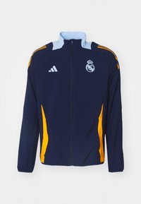 Blå atletisk jakke med lyseblå krage, gule sidelommer, Adidas-logo og brodert lagmerke. Laget av lett stoff.