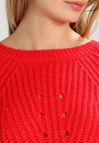 Pull en tricot rouge avec un col large, une texture côtelée et des motifs de trous espacés uniformément sur tout le tissu.