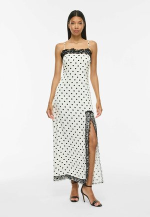 Femme portant une robe longue blanche sans manches à pois noirs, bordure en dentelle et fente haute sur la cuisse, associée à des sandales noires à talons hauts.