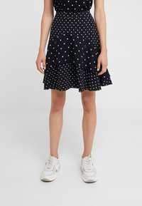 Jupe noire à pois blancs, avec un design superposé et des volants à l'ourlet. Associée à des baskets blanches.