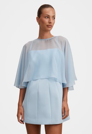 Nő kontyba fogott hajjal, áttetsző világoskék köpenyt visel, amely egy hozzáillő, ujjatlan ruha felett van, redős szoknyával, szürke háttér előtt áll.