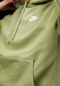 Sudadera verde de tela suave con un bolsillo frontal, capucha con cordón y un logo blanco de Nike en el pecho. Textura lisa.