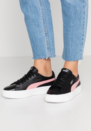 Schwarze Plateau-Sneaker mit pinkem Puma-Streifen, getragen zu gekürzten blauen Jeans auf hellem Fußboden.