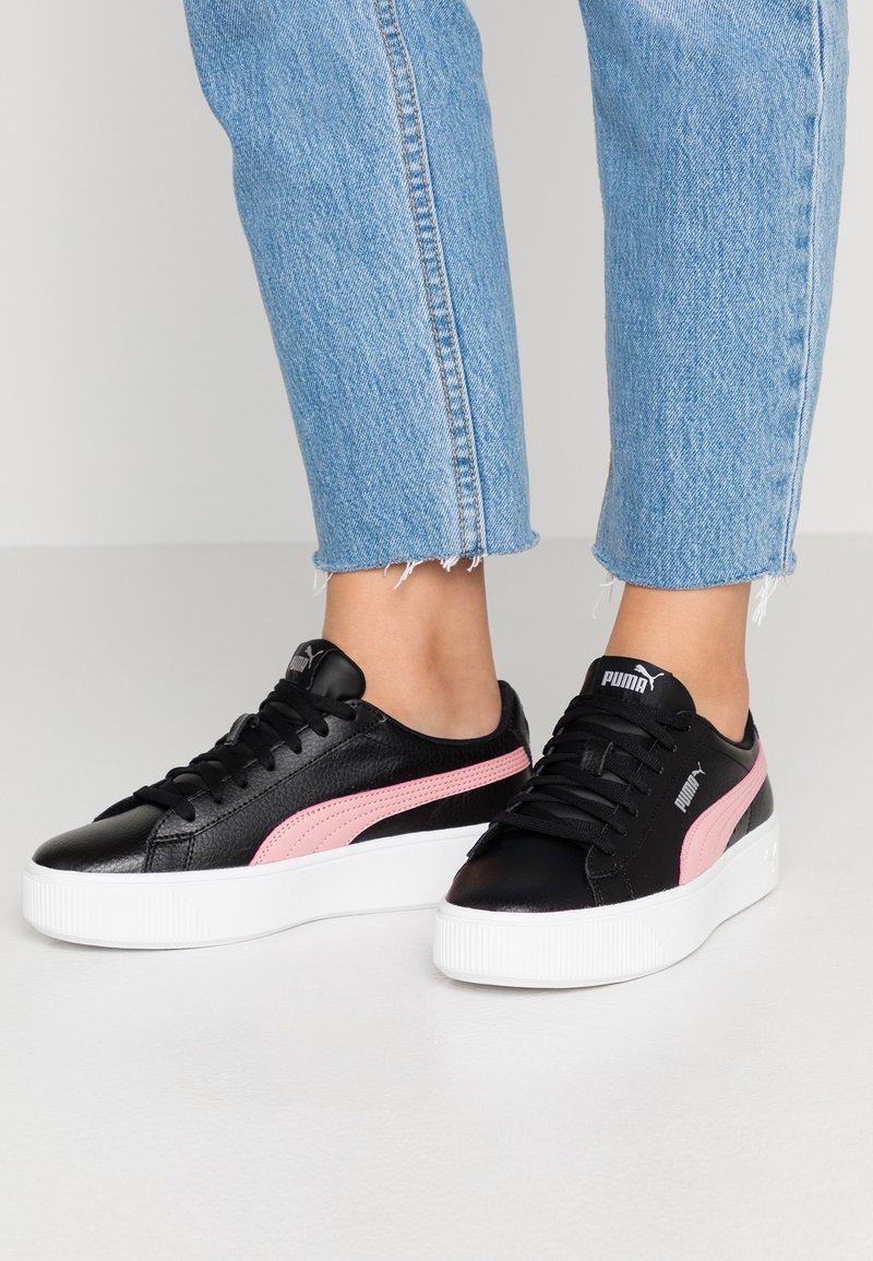 Baskets à plateforme noires avec bande rose Puma portées avec un jean bleu raccourci sur un fond de sol clair.