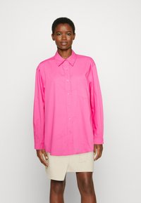 Camisa de manga longa rosa em algodão, com colarinho clássico, fecho de botão e um bolso no peito. O tecido possui uma textura suave.