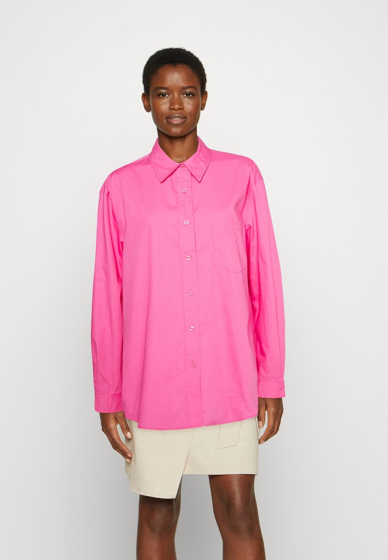 Camisa de manga longa rosa em algodão, com colarinho clássico, fecho de botão e um bolso no peito. O tecido possui uma textura suave.