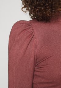 Pull bordeaux en tissu côtelé texturé. Comprend des détails d'épaules bouffantes et une silhouette ajustée. Finition lisse avec un léger éclat.