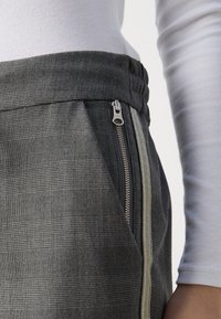 Grå rutig byxor med en strukturerad tyg, som har en sidodragkedja och ett kontrasterande ljust rand längs sömmen. Midjebandet är elastiskt.