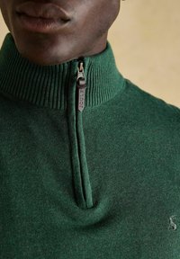 Artimo plano nuotrauka, kurioje matyti žmogus, vilkintis tamsiai žalią megztinį su puse užtrauktuko ir mažą siuvinėtą logotipą ant krūtinės.