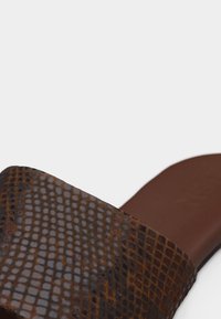 Sandale à glissière marron avec bride en cuir à motif quadrillé texturé et semelle intérieure lisse sur un fond clair.