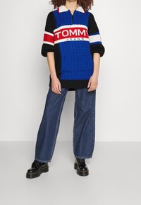 Pull-over tricoté bleu et noir avec un col zippé, présentant une bande blanche et rouge ainsi que le logo "TOMMY JEANS". Associé à un jean décontracté et des bottes noires.