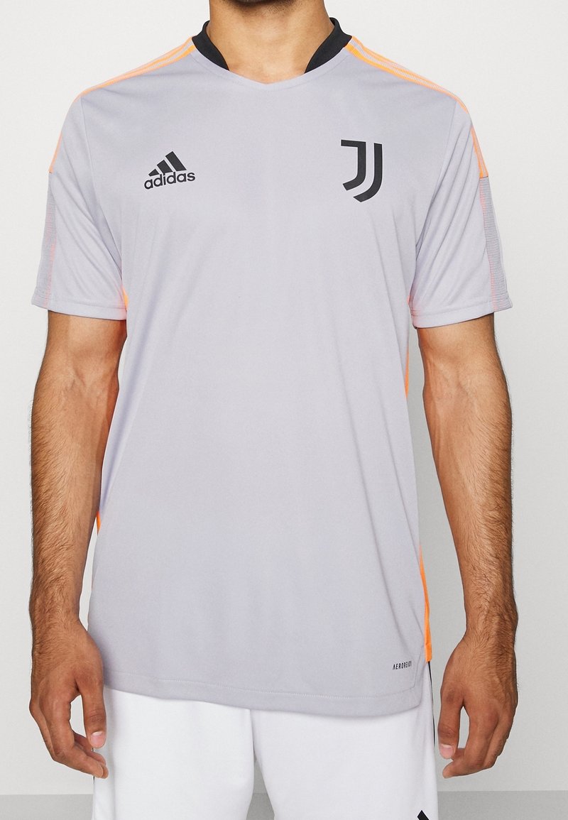Jersey deportivo de manga corta en gris claro con cuello negro y logo de Adidas, con acentos naranja en los hombros y los lados.