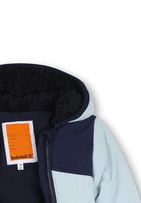 Blauwe en lichtblauwe gevoerde hoodie-jack met een zachte voering, gewatteerde textuur en een zichtbaar merketiket in oranje en wit binnen de kraag.