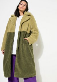 Cappotto in pile verde oliva e verde chiaro con colletto, vestibilità oversize, tasche laterali e una texture morbida, che arriva fino a metà coscia.