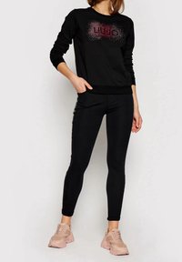 Zwarte sweatshirt met sprankelende "LIU JO" letters, gecombineerd met zwarte legging. Model draagt roze sportieve sneakers, wat een casual outfit laat zien.