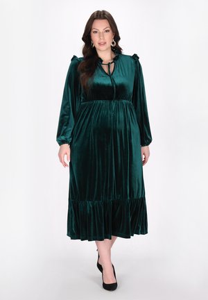 Lungo abito in velluto verde smeraldo con maniche lunghe, dettagli arricciati sulle spalle, scollatura legata e orlo a balze stratificate.