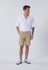 White short-sleeve polo with a V-neck; paired with beige shorts; model wears gray and black sneakers, standing against a white brick background.