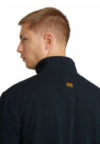 Felpa color blu navy con collo alto, realizzata in tessuto a coste, con un'etichetta quadrata sul retro vicino al collo.
