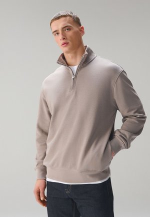 Giovane uomo che indossa una felpa beige con mezza cerniera sopra una camicia bianca e jeans scuri, in piedi su uno sfondo grigio chiaro uniforme.