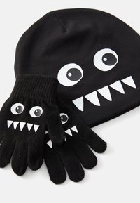 Zwarte gebreide beanie en handschoenen met grote witte ogen en grijnzende mondjes, gemaakt van zacht materiaal met ribbels aan de manchet voor extra warmte.