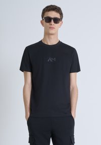 Camiseta negra de algodón con cuello redondo, que presenta un sutil logo en relieve en el pecho. Complementada con pantalones negros a juego y gafas de sol.