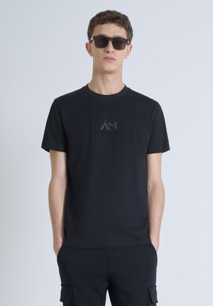 Black t-shirt made of cotton with a round neckline, featuring a subtle embossed logo on the chest. Complemented by matching black pants and sunglasses.