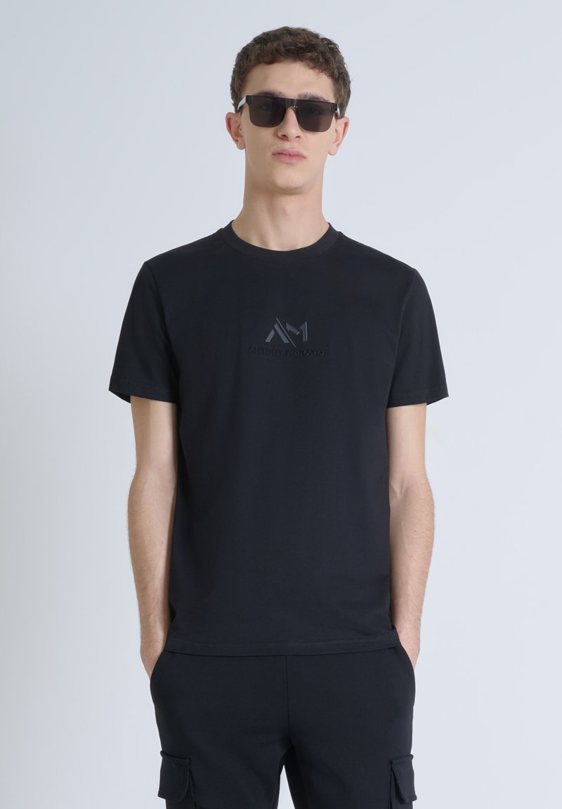Camiseta negra de algodón con cuello redondo, que presenta un sutil logo en relieve en el pecho. Complementada con pantalones negros a juego y gafas de sol.