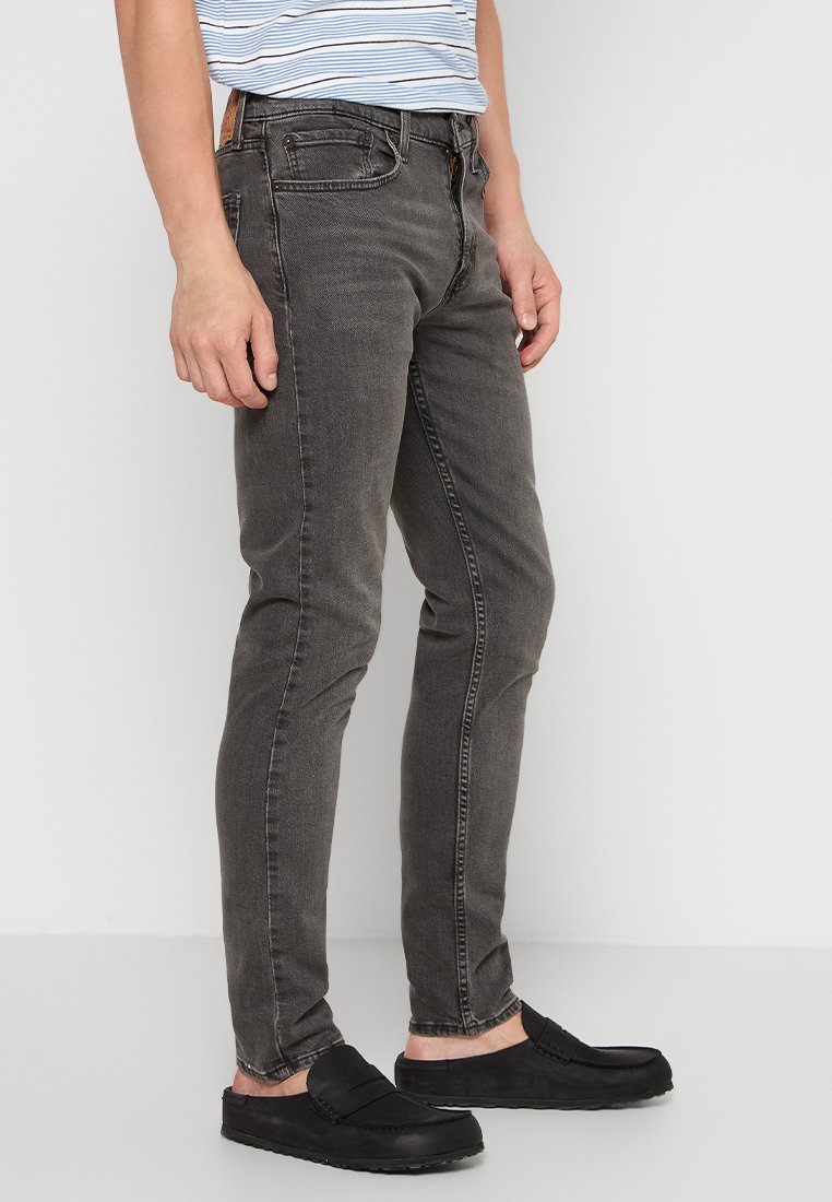 Schmal geschnittene, dunkelgraue Jeans aus Denim mit klassischem Fünf-Taschen-Design, subtilen Abnutzungserscheinungen und einem Reißverschluss. Getragen mit schwarzen Slippern.