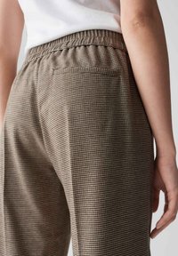 Bruine geruite broek met elastische tailleband, voorzien van een achterzak en een textuurstof. Close-up van het achterontwerp.