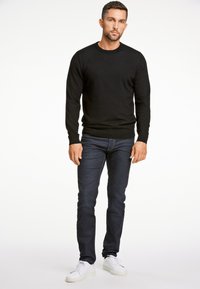 Czarny sweter z długim rękawem, ciemnoniebieskie dżinsy i białe trampki. Model stoi z rękami swobodnie po bokach na tle jednolitego tła.