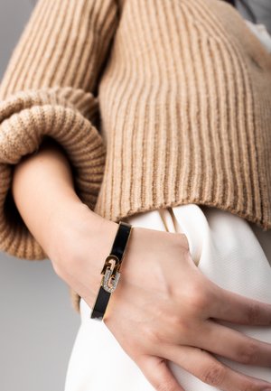 Black glossy bracelet with gold accents and diamond-like embellishments, worn on a hand against a backdrop of a ribbed beige sweater.