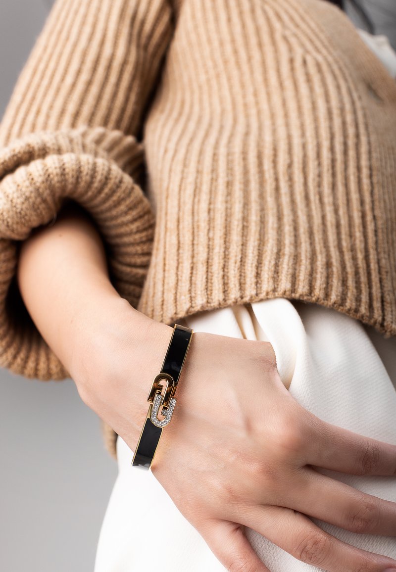 Black glossy bracelet with gold accents and diamond-like embellishments, worn on a hand against a backdrop of a ribbed beige sweater.