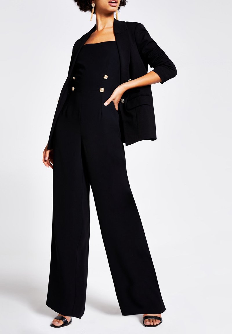 Femme portant une combinaison noire sans manches avec des boutons dorés et un blazer noir assorti, debout avec une main sur la hanche.