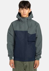 Giacca con cappuccio di colore grigio-verde nella parte superiore e blu navy nella parte inferiore. Presenta una tasca frontale, chiusura con zip e polsini elasticizzati.